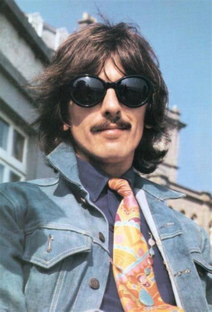 George Harrison wearing rectangular sunglasses, turtleneck, sport coat, and hat while waving to the camera as he boards a plane.