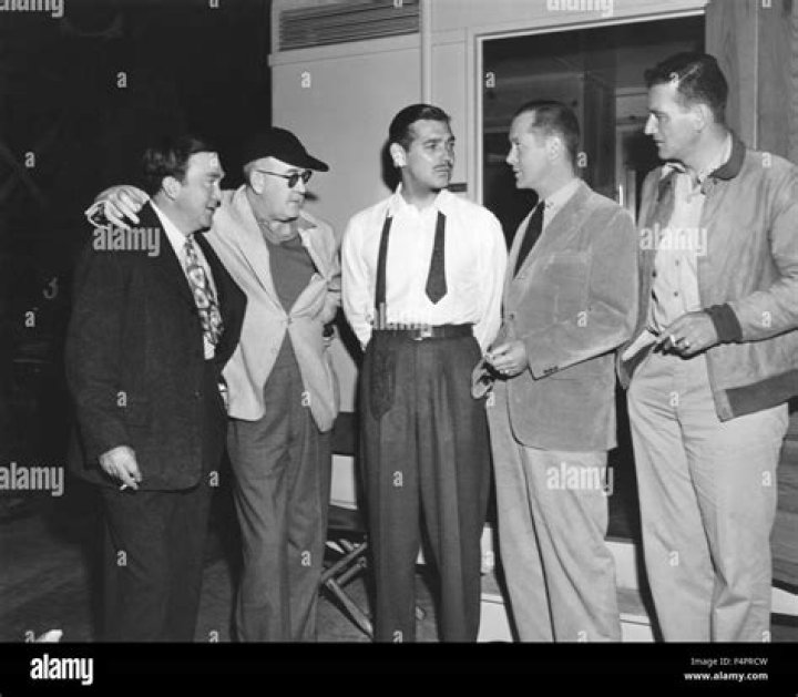 Thomas Mitchell, John Ford, Clark Gable, Robert Montgomery, and John Wayne standing around on the set wearing suits and smoking cigarettes